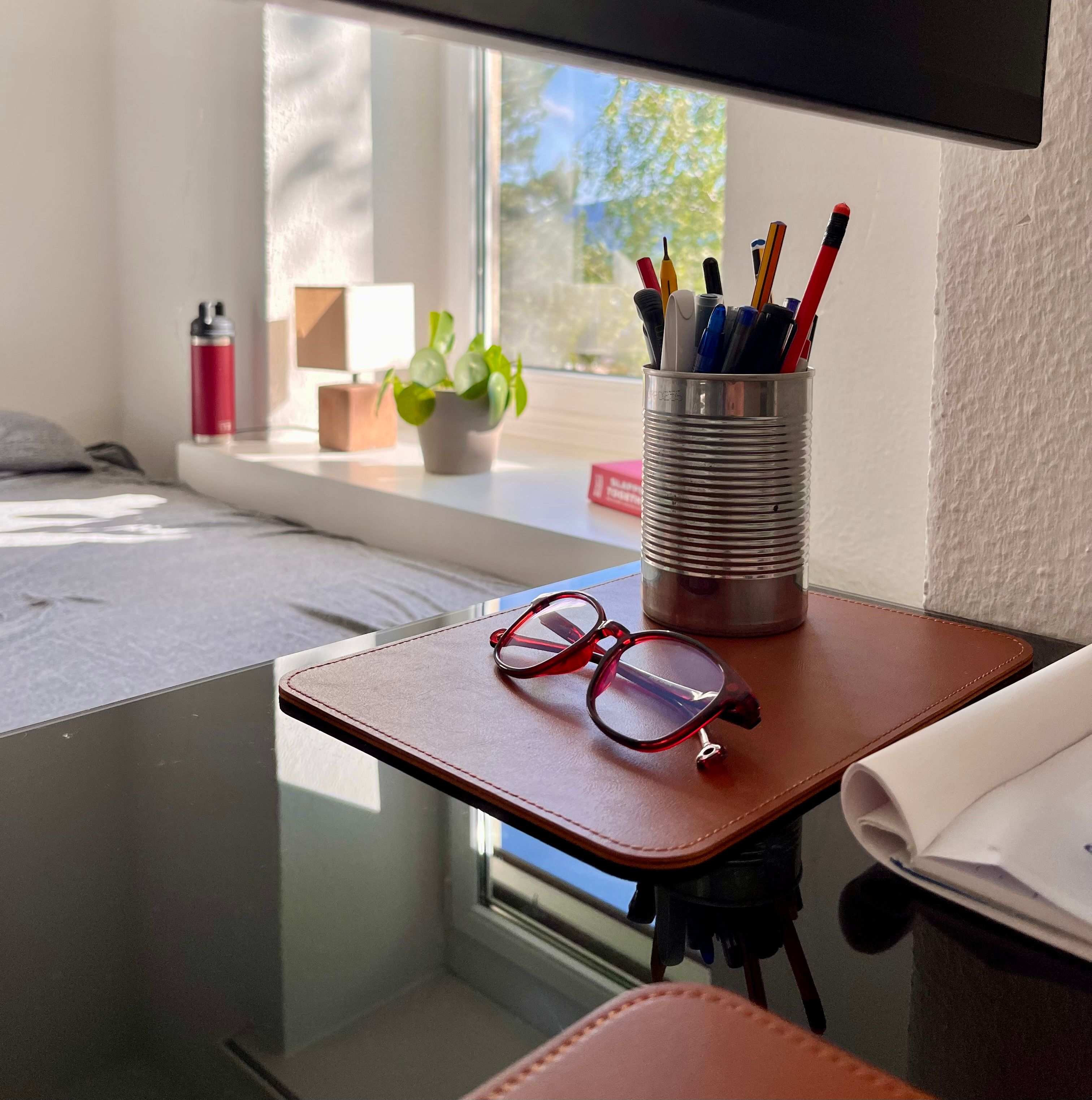 pens in penholder, a pair of glasses on a deskmat, in the background a window with a lamp, a plant, a book and a metal water bottle